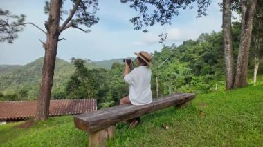 Asyalı bayan turist dışarıda, yemyeşil bir alanda bir parkta kamerayla fotoğraf çekiyor. Yaz turizmi kavramı.