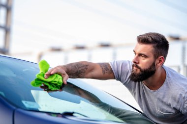 Genç bir adam gün ışığında dışarıda bir bezle arabasını temizliyor..