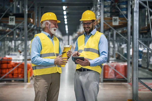 Engineers walking through large warehouse center. They are checking shelves full with boxes and products. Warehouse workers using digital tablet.