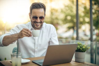 Mutlu iş adamı bir kafede oturuyor, kahve içiyor ve dizüstü bilgisayarda bir web sitesi izliyor.. 