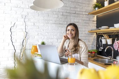 Evde dizüstü bilgisayarla çalışırken gülümseyen genç bir kadın kahvaltı yapıyor..