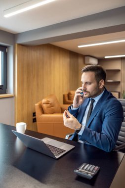 Genç CEO, ofisini akıllı telefon ve dizüstü bilgisayar ile kullanıyor. İş adamı istatistiklerle çalışıyor, dizüstü bilgisayara bakıyor ve akıllı telefondan konuşuyor..