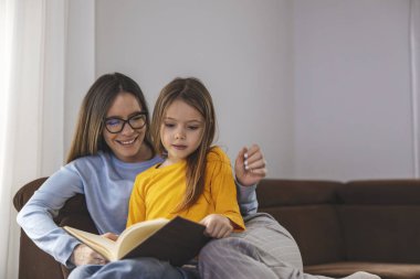 Mutlu anne ve kız gülümseyip kitap okurken hafta sonu evde kanepede dinleniyorlar..