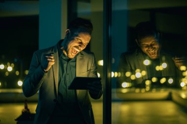Ofisinde duran gülümseyen genç işadamı ve arka planda şehir ışıklarıyla gecenin geç saatlerinde dijital tabletle çalışıyor. Başarıyı kutlayın..