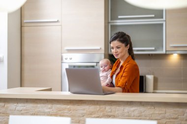 Genç anne mutfak ofisinde dizüstü bilgisayarı ve bebeğiyle. Ev kavramından çalış.