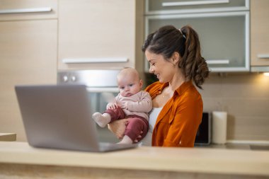 Genç anne mutfak ofisinde dizüstü bilgisayarı ve bebeğiyle. Ev kavramından çalış.