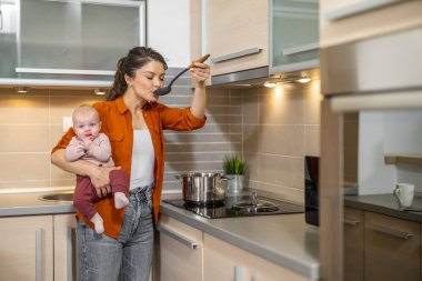 Genç anne bebekle mutfakta, elinde bebekle öğle yemeği hazırlıyor. Birlikte mutlu anlar.