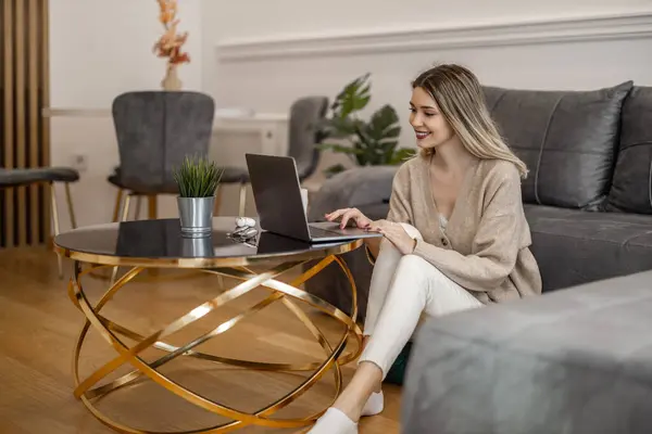 在家里工作时 用笔记本电脑来吸引穿着休闲装的年轻女性 在家里工作的自由职业者微笑 图库图片