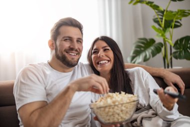 Young happy couple watching movie on TV and eating popcorn while relaxing in the living room.