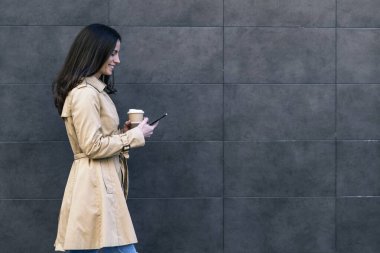 Elinde kahve tutan çekici bir kız, elinde akıllı bir telefonla şehirde yürüyor..