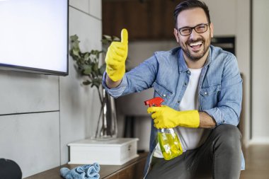 Sarı lastik eldivenli genç adam sprey ve giysi kullanarak toz temizliyor. Ev işi, temizlik ve insanlar konsepti..