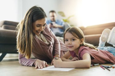 Genç anne ve küçük kızı birlikte oynuyorlar. Renkli kalemlerle aile çizimi.