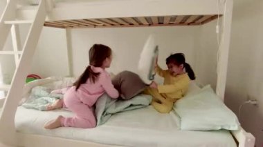 Sevimli küçük kızlar evde, yatakta yastık savaşı yapıyorlar. Mutlu kız kardeşler yastıklarla savaşıyor..
