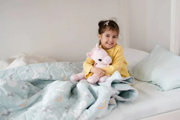Morning awakening little child girl with her toy in bed. Happy girl in pajamas sitting on bed right after awaking. Happy family concept