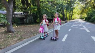 Sırt çantalı iki kız parkta scooter kullanıyor. Çocuklar dışarıda mobiletlerle eğleniyor..