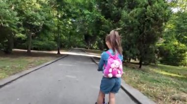 Pembe sırt çantalı şirin bir kız parkta scooter kullanıyor.