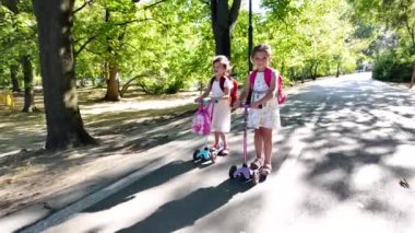 Sırt çantalı iki kız parkta scooter kullanıyor. Çocuklar dışarıda mobiletlerle eğleniyor..