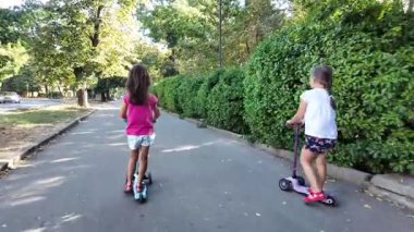 Parkta scooter süren iki kız. Çocuklar dışarıda mobiletlerle eğleniyor.. 