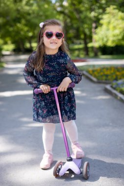 Kalp şeklinde güneş gözlüğü takan gülümseyen kız parkta scooter sürüyor. Açık havada güneşli bir günün tadını çıkarıyor, çocukluk eğlencesini, mutluluğu ve aktif yaşamı temsil ediyor.
