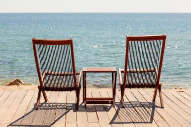 Sunny Beachfront güvertesinde iki tahta şezlong ve denize bakan masa. Rahatlama, Yaz Tatili, Romantik Kaçış Konsepti