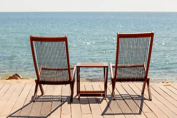 Sunny Beachfront güvertesinde iki tahta şezlong ve denize bakan masa. Rahatlama, Yaz Tatili, Romantik Kaçış Konsepti
