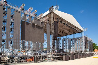 Açık havada LED ekranlar, aydınlatma platformları ve açık mavi gökyüzünün altında kumlu bir plaja kurulmuş ses sistemleri ile büyük bir konser sahnesi inşa ediliyor.