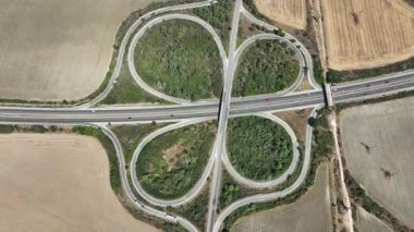 Simetrik yonca yaprağı otoyol kavşağının yukarıdan aşağı görüntüsü. Yol ağındaki otomobiller kırsal bir alanda ulaşımı ve lojistiği gösteriyor, bağlantı ve altyapıyı sembolize ediyor.