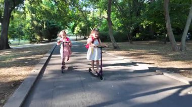 Sırt çantalı iki kız parkta scooter kullanıyor. Mutlu çocuklar dışarıda eğlenmeyi, arkadaşlığı ve aktif bir çocukluk hayatı sürmeyi seviyorlar..