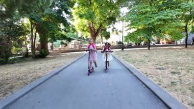 Sırt çantalı iki kız parkta scooter kullanıyor. Mutlu çocuklar dışarıda eğlenmeyi, arkadaşlığı ve aktif bir çocukluk hayatı sürmeyi seviyorlar..