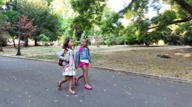 Mutlu çocuklar. Okul çantalı iki kız parkta yürüyor. İki küçük kız kardeş birlikte yürüyorlar, konuşuyorlar. En iyi arkadaşlar