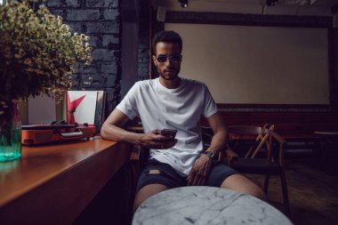 Beyaz tişörtlü Afro-Amerikalı bir adam bir kafede oturur ve telefonu kullanır. Model.