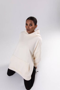 Attractive african american woman in oversized white hoodie sits on white background. Mock-up..