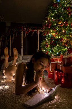 İç çamaşırlı güzel, seksi esmer bir kadın yalan söylüyor ve elinde bir Noel ağacının arka planına karşı bir hediye tutuyor. Yüksek kalite fotoğraf