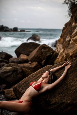 Bir kadın kayalık kıyılarda huzurlu ve huzurlu bir anın tadını çıkarır. Parlak kırmızı bir bikini giyerken zarifçe poz verir.