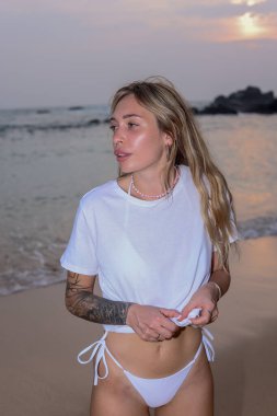 Güzel bir günbatımında zarif beyaz mayo giymiş bir kadının yer aldığı huzurlu ve huzurlu bir sahil sahnesi.