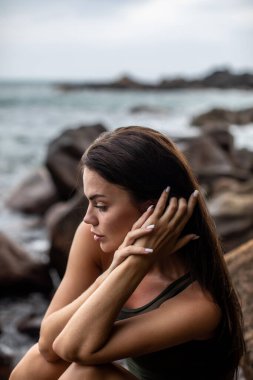 Düşünceli bir kadın sakin bir sahil şeridinde kayaların üzerinde oturmuş güzelliğin özünü bütünüyle somutlaştırıyor.