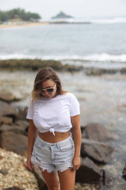 Bir kadın, köpüklü suyun yanındaki büyük bir kayanın üzerinde kendine güvenerek duruyor, şık beyaz bir bluz ve günlük şort giyiyor.