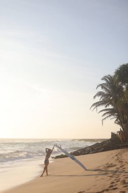 Bir insan güzel bir sahilde elinde sörf tahtasıyla duruyor, üzerinde güneş ışıldarken bazı dalgaları yakalamaya hazır.