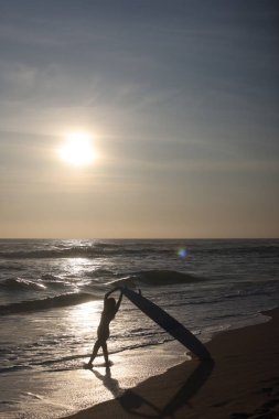 Güzel, sakin bir gün batımı manzarası dalgaların yanında sörf tahtası tutan bir sörfçünün siluetini gösteriyor.
