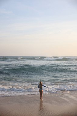Bir kadın, elinde sörf tahtası, suda eğlence ve heyecan dolu bir güne hazır bir şekilde güzel plajda rahatça yürüyor.