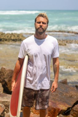 Bir sörfçü renkli tahtasıyla güvenle poz verir kayalık bir plajda, okyanusta bazı dalgaları yakalamak için tamamen hazırdır.