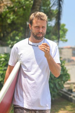 Bir adam canlı bir tropikal ortamda sörf tahtasıyla durur güneş gözlüğü takar ve heyecan verici bir maceraya hazırdır.