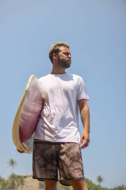 Bir sörfçü, parlak mavi bir gökyüzünün altında canlı bir geçmişe dönük sörf tahtasıyla kendinden emin bir şekilde durur ve neşe saçar.