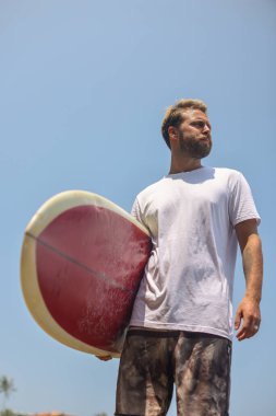 Bir sörfçü, parlak mavi bir gökyüzünün altında canlı bir geçmişe dönük sörf tahtasıyla kendinden emin bir şekilde durur ve neşe saçar.