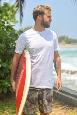 Kendinden emin bir sörfçü, güneşli bir günde okyanus dalgalarının yanında sörf tahtasıyla gururla durur.