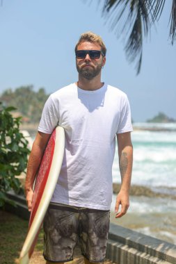 Kendinden emin bir sörfçü, güneşli bir günde okyanus dalgalarının yanında sörf tahtasıyla gururla durur.