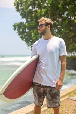 Güneşli bir gökyüzünün altında sörf tahtası taşıyan bir sörfçü kayalık kıyılarda yürüyor.