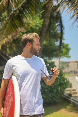 Bir adam canlı bir tropikal ortamda sörf tahtasıyla durur güneş gözlüğü takar ve heyecan verici bir maceraya hazırdır.