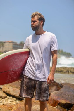 Bir sörfçü, parlak mavi bir gökyüzünün altında canlı bir geçmişe dönük sörf tahtasıyla kendinden emin bir şekilde durur ve neşe saçar.