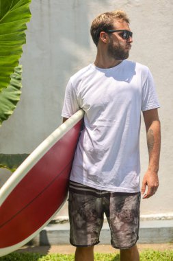 Kendinden emin genç bir adam gururla ayakta duruyor. Dalgalı ve güneşli bir tropikal plaj yerinde.